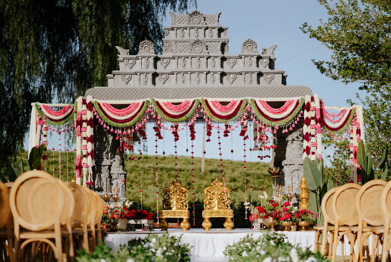Temple-style mandap inspiration: carved gray pillars, a tiered temple roof, pink, white and green garlands, and gold ceremony chairs